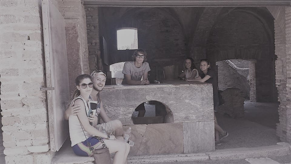 Photograph of five students standing in a Pompeiian shop. One student stands behind the counter. Two students stand on the other side of the counter. Two more students sit on a bench close to the road.